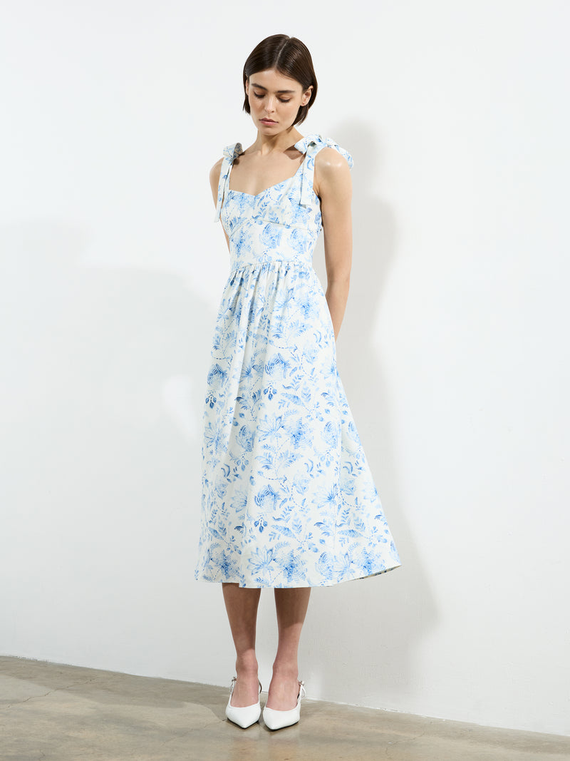 A woman stands indoors against a plain white wall, looking down with hands clasped behind her back, wearing the French Connection Serenity Dale Crepe Midi Dress in light blue botanical print and white pointed-toe heels.